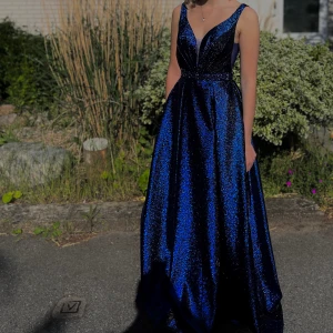 Blå glittrig långklänning - Säljer en fantastisk blå glittrig långklänning perfekt för fest eller nyår. Klänningen har en djup v-ringning och är ärmlös. Materialet är glittrande och fångar ljuset vackert!