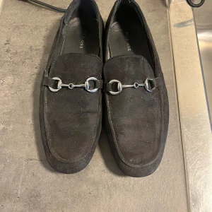 Svarta mockaloafers från Pier One - Snygga svarta loafers i mocka från Pier One. De har en stilren design med metallspänne på ovansidan. Perfekta för både vardag och festliga tillfällen. Skorna är i bra skick och redo att användas direkt.