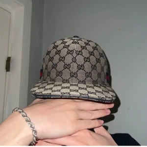 Beige keps från Gucci - Säljer en snygg beige keps från Gucci med det klassiska GG-mönstret. Kepsen har en justerbar rem baktill och detaljer i rött och blått på sidan. Perfekt för att ge din outfit en lyxig touch!