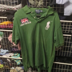  Ralph Lauren - Säljer en grön pikétröja från Ralph Lauren med 'Great Britain' och flaggor broderade på bröstet och ryggen. Tröjan har en klassisk krage och knappar framtill. Perfekt för en avslappnad stil eller en dag på stan. 🏇storlek xxl men sitter som L, solblek på kragen som man ser på bild 