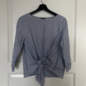 Randig blus från Lindex - Säljer en superfin randig blus från Lindex i storlek S. Den har blå och vita ränder och en snygg knytdetalj framtill. Blusen har trekvartsärmar och är perfekt för både vardag och fest. Passar bra till jeans eller kjol!