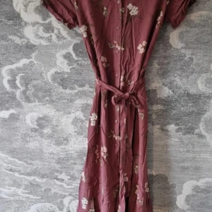 Blommig klänning i viskos - Säljer en superfin blommig klänning i 100% viskos. Den är i en härlig mörkrosa färg med söta blommor och fjärilar. Klänningen har korta ärmar och knappar framtill, samt ett knytband i midjan för en smickrande passform. Perfekt för vår och sommar!