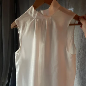 Vit ärmlös blus med knytband - Säljer en elegant vit blus med knytband vid nacken. Den är ärmlös och har en lös passform, perfekt för både vardag och fest. Blusen är i nytt skick och ger en stilren look.