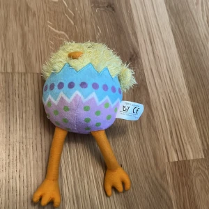 Färgglad påskkyckling i polyester - En söt och färgglad påskkyckling med mjuk, fluffig gul päls på huvudet och ett mönstrat kropp i lila, blått och grönt. Tillverkad i 100% polyester, perfekt som dekoration eller leksak under påsken.