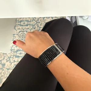 Svart glittrigt armband - Snygg svart armband (cuff) med glittriga detaljer. Klockan har en bred design med ett silverfärgat spänne och svarta stenar. Perfekt för att ge en elegant touch till din outfit.