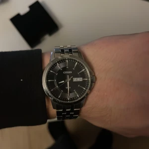 Citizen herrklocka i silver - Snygg Citizen herrklocka med svart urtavla och datumvisning. Klockan har en stilren design med en silverfärgad länkarmband och tydliga markeringar på urtavlan. Perfekt för den som gillar en klassisk look. Köpt för 1000 kr. Vet inte måttet.