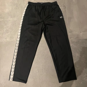 Svarta byxor från Fred Perry - Säljer ett par svarta byxor från Fred Perry i mycket bra skick. De har en elastisk midja och vita detaljer längs sidorna med Fred Perry-loggan. Perfekta för en avslappnad stil eller träning. Tillverkade i Portugal. Byxorna är passar för M/L