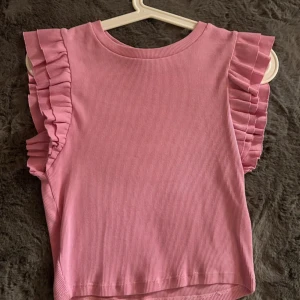 Rosa ärmlös topp med volanger - Säljer en supersöt rosa ärmlös topp med volanger på axlarna. Toppen är ribbad och har en croppad passform, perfekt för sommardagar eller en festlig look. Den är i mycket bra skick och redo för nya äventyr!