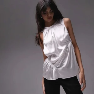 Vit ärmlös topp från Topshop - Snygg vit ärmlös topp från Topshop i ett glansigt material. Toppen har en rund halsringning och ett elastiskt band i midjan för en smickrande passform. Perfekt för en stilren look.