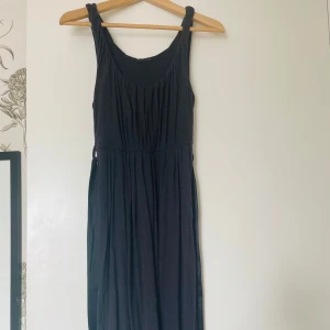 Svart klänning från Bruuns Bazaar - Elegant svart klänning från Bruuns Bazaar i mycket bra skick. Den har en stilren design med ett bälte i midjan för en smickrande passform. Mjuk och bekväm. Går nedanför knäna. Perfekt för både fest och mer formella tillfällen. Klänningen är ärmlös och har en klassisk, tidlös stil.