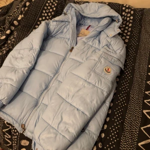 Moncler Puffer Jacka - Säljer en snygg ljusblå dunjacka med märket Moncler jackan har en huva och dragkedja. Jackan har ett broderat Moncler-märke på ärmen och en unik serietidningsetikett på insidan. Perfekt för kyliga dagar! Och perfekt för dig som har stil. Ställ fler frågor privat!!