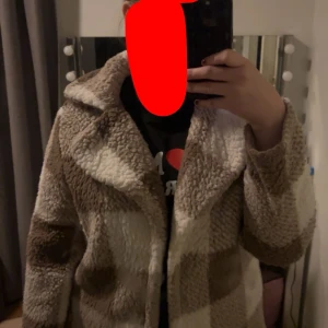Rutig teddyjacka i beige och brun - Mysig teddyjacka med rutigt mönster i beige och bruna nyanser. Jackan har en klassisk krage och långa ärmar. Den är ganska tunn så jag rekommenderar att ta den på hösten eller våren. Använde den i typ en vecka 2022. så den är fortfarande ganska fluffig. Lappen är sönder. Jag vet inte vilket varumärke jag köpte den ifrån, men tror att den är från SHEIN.