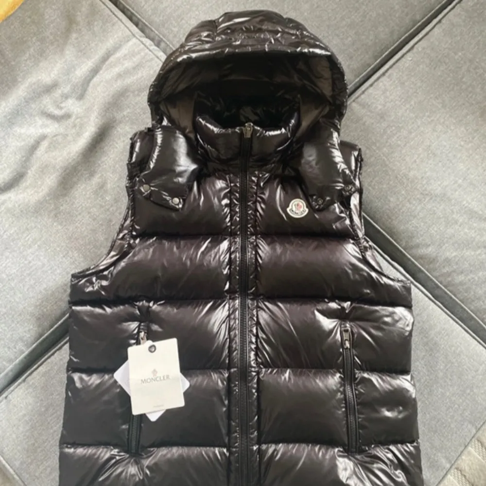 Säljer en snygg svart dunväst från Moncler i nytt skick. Västen har en glansig finish och är quiltad med dragkedja framtill. Den har en praktisk huva och två fickor med dragkedja. Perfekt för höst och vinter!. Jackor.