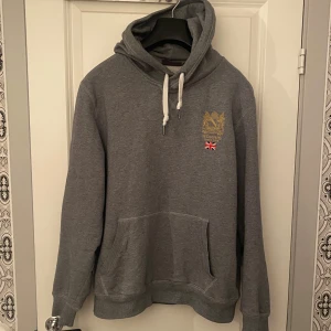 Grå hoodie från Morris - Säljer en snygg grå hoodie från Morris med broderad logga och brittisk flagga på bröstet. Den har en klassisk känguruficka och justerbar huva med snören.   Jag är 184cm lång och väger ca 75kg. Vid minsta fråga eller fundering hör av er🤝