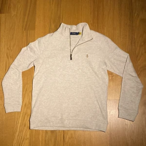Beige tröja från Polo Ralph Lauren - Snygg beige tröja från Polo Ralph Lauren med dragkedja vid halsen och broderad logga på bröstet. Perfekt för en stilren och avslappnad look.
