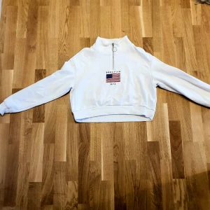 Vit tröja med dragkedja från Divided - Säljer en vit tröja från Divided med halv dragkedja och tryck av amerikansk flagga samt texten 'Brooklyn NYC'. Tröjan har långa ärmar och en croppad passform. Perfekt för en avslappnad stil!