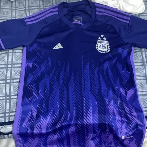 Lila Argentina fotbollströja från Adidas - Säljer en snygg lila fotbollströja från Adidas med argentina logga på bröstet. Tröjan är kortärmad och har ett coolt mönster i olika nyanser av lila. Den är tillverkad i Aeroready-material som håller dig sval och torr. Perfekt för både träning och match, även för dig som vill se bra ut i den!🤩