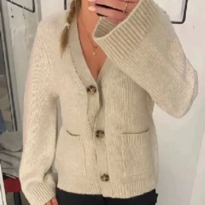 Beige stickad kofta - Säljer en supermysig beige stickad kofta med stora knappar framtill. Perfekt för kyliga dagar och har två praktiska fickor. Den är långärmad och har en skön, ribbad kant vid ärmslut och nederkant. Passar perfekt till både vardag och lite finare tillfällen. 