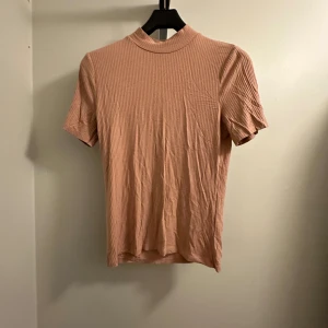 Beige/laxfärgad ribbad t-shirt - En snygg beige/laxfärgad ribbad t-shirt i storlek M från New Yorker. Den har en normal passform och korta ärmar. T-shirten är enkel men stilren, vilket gör den lätt att matcha med olika outfits. Oanvänd!