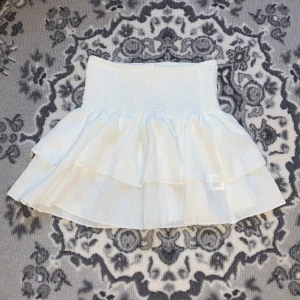 Vit volangkjol - Superfin vit kortkjol med volanger, perfekt för sommaren! Den har en elastisk midja som gör den bekväm och lätt att bära. Kjolen är i ett luftigt material och längden på kjolen är 34 cm. Har bara använt 1-2 gånger 🤗