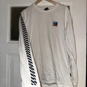 Sweeet STBDS x Helly Hansen tröja  - Säljer en vit långärmad tröja från Helly Hansen med coola svarta ränder längs ärmarna. Tröjan har en stor logga på bröstet och ryggen. Perfekt för en avslappnad stil eller för att sticka ut lite extra. Tillverkad i bomull för en skön känsla. Passar perfekt till både vardag och chill.