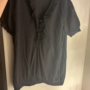 Svart blus från H&M - Säljer en svart blus från H&M med korta ärmar och dekorativa detaljer vid halsringningen. Perfekt för en stilren look.