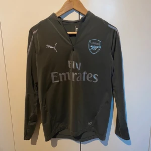 Grå långärmad Arsenal fotbollströja  - Säljer en grå långärmad fotbollströja från Puma. Tröjan har en stilren design med klubbmärke på bröstet och är i mycket bra skick. Perfekt för träning eller som supportertröja. Passar bra för kyligare dagar på planen. 
