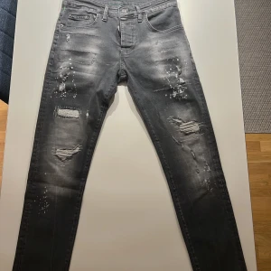 Grå jeans från Dsquared2 - Säljer ett par grå Dsquared2 jeans i slim fit. De har en cool distressed look med slitningar och färgstänk. Perfekta för en avslappnad men stilren outfit. Midjan är normal och de har klassiska fem fickor. Passar perfekt för höst och vår!