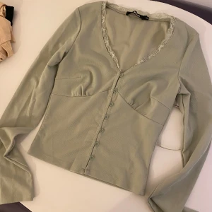 Grön långärmad topp med spetsdetaljer - Snygg grön topp med långa ärmar och knappar framtill. Den har en v-ringad design med spetsdetaljer längs halsringningen. Använd endast 1 gång, passar både XS och S