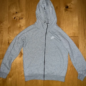 Grå hoodie från Nike - Säljer en grå hoodie från Nike i bra skick. Den har en dragkedja framtill och en klassisk Nike-logga på bröstet. Perfekt för både träning och vardag. Den är superbekväm och har en normal passform. Huvan är justerbar med snören. Passar perfekt till höst och vår!