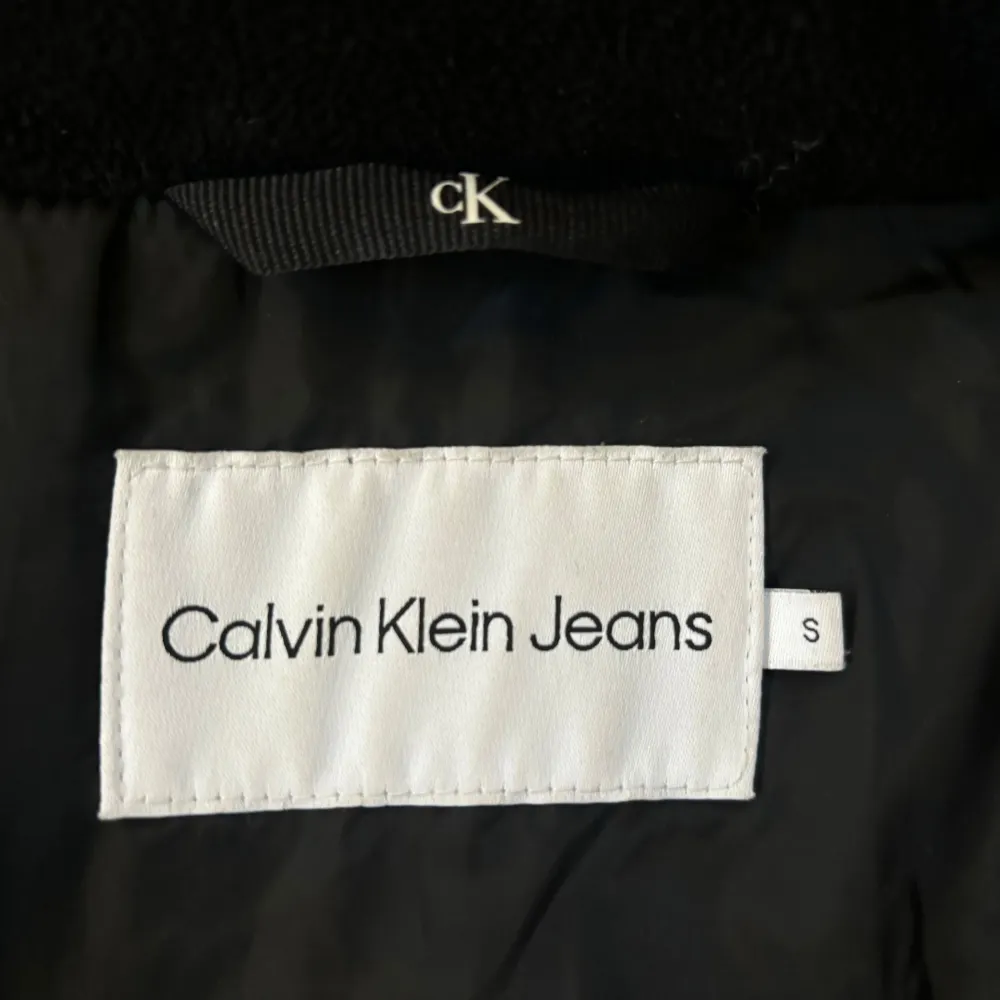 Säljer en snygg svart pufferjacka från Calvin Klein Jeans i glansigt material. Jackan har en stor huva och dragkedja framtill. Perfekt för kyliga dagar och ger en stilren look. Passar både till vardags och festliga tillfällen.. Jackor.