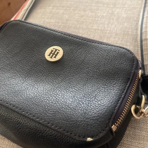 Mörk blå axelväska från Tommy Hilfiger - Snygg mörk blå axelväska i skinn från Tommy Hilfiger😍😍 med guldfärgade detaljer och dragkedja. Väskan har en justerbar axelrem med röd och vit randig design. Perfekt för att bära dina nödvändigheter med stil. Dragkedjan där fram har gått av men går fortfarande att använda som vilken annan💕💕