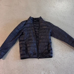 Svart pufferjacka från Emporio Armani - Snygg svart pufferjacka från Emporio Armani EA7 med quiltad design. Jackan har en dragkedja framtill och är perfekt för kyligare dagar. Den har en stilren look med märkets logga på bröstet.