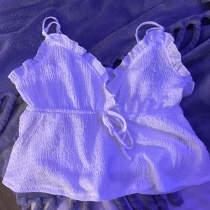 Vit ribbad topp med volangdetaljer - Säljer en söt vit ribbad topp med volangdetaljer och knytning framtill. Toppen har smala axelband och en feminin design som passar perfekt för sommardagar.