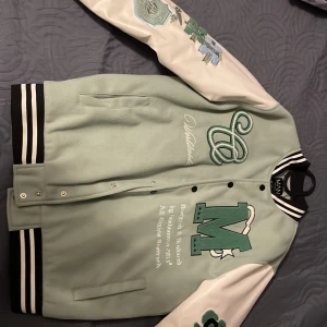 Grön och vit bomberjacka från boohooMAN - Snygg grön och vit bomberjacka från boohooMAN med broderade detaljer och stora bokstäver. Jackan har knappar framtill och randiga muddar vid krage, ärmslut och nederkant. Perfekt för en cool och avslappnad stil.