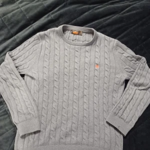 Ljusblå stickad tröja Morris - Säljer en stilren grå stickad tröja från Morris med klassiskt flätmönster. Tröjan har en liten orange broderad detalj på bröstet och ribbade muddar vid ärmslut och nederkant. Perfekt för kyliga dagar! OBS MÖRKARE I VERKLIGENHETEN