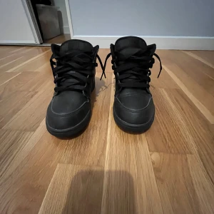 Svarta Nike Air Jordans - Snygga svarta Nike Air Jordans med klassisk design och snörning. Perfekta för en stilren look med en sportig touch. Skorna har en diskret logga på sidan och är tillverkade i syntetmaterial.