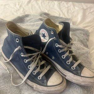 Blå Converse All Star sneakers - Säljer ett par klassiska blå Converse All Star sneakers i bra skick. De har den ikoniska vita sulan och snörning, med det välkända märket på sidan. Perfekta för en avslappnad stil och funkar året runt. Lite slitna men fortfarande snygga och bekväma!