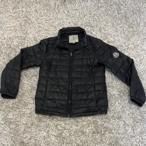 Moncler damjacka - Säljer en snygg svart dunjacka från Moncler i mycket bra skick. Jackan har en quiltad design med dragkedja framtill och en klassisk krage. Perfekt för höst och vinter, håller dig varm och stilren. Moncler-logga på ärmen ger en exklusiv touch. Passar både till vardags och lite finare tillfällen.