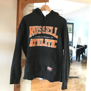 Svart hoodie från Russell Athletic - Säljer en svart/grå hoodie från Russell Athletic i storlek medium. Den har en stor, orange och vit logga på framsidan och en klassisk känguruficka. Perfekt för en avslappnad stil eller träning. Huvan har dragsko för justering. Skön och bekväm för kyliga dagar.
