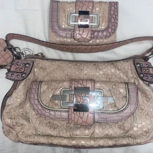Beige och rosa väska och plånbok från Guess - Säljer en snygg beige axelväska och matchande plånbok från Guess. Väskan har ett glittrigt mönster med rosa detaljer och en stor metallspänne framtill. Plånboken har samma stil med krokodilmönstrade detaljer. Perfekt för en chic look!