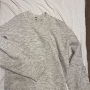 Grå stickad tröja - Mysig grå stickad tröja i ett mjukt material. Perfekt för kyliga dagar och att mysa i hemma. Tröjan har en klassisk rund hals och långa ärmar med ribbade muddar. Passar bra till både jeans och leggings för en avslappnad look.