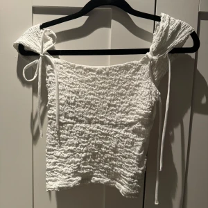 Vit smockad topp med knytband - Säljer en superfin vit smockad topp med knytband på axlarna. Perfekt för sommardagar eller en festlig kväll. Toppen är i ett stretchigt material och har en söt, feminin stil. Passar perfekt till både jeans och kjol!