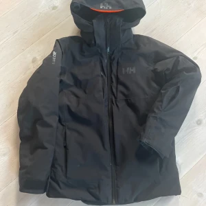 Svart skidjacka JR Alpha från Helly Hansen - Säljer en svart vinterjacka från Helly Hansen i nytt skick. Jackan har fickor för liftkort, hjälmanpassad huva som är avtagbar, snölås och är perfekt för kalla dagar i backen. Den är utrustad med RECCO-teknologi och Primaloft-isolering för extra värme och säkerhet. Perfekt för vintersport eller vardagsbruk. Storlek: JR 140 (10). Nypris: 2200 kr 