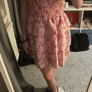 Rosa blommig klänning med volanger - Charmig rosa klänning med blommigt mönster och volanger. Axelbandslös design med knytning framtill och smockad midja för en bekväm passform. Perfekt för sommarens varma dagar. Använd 1 gång, skriv för mer information💕