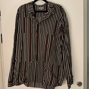 Randig skjorta från COS - Säljer en stilren randig skjorta från COS i storlek XL. Skjortan har en klassisk krage och är långärmad, perfekt för både vardag och fest. Färgerna är svart, vit och röd