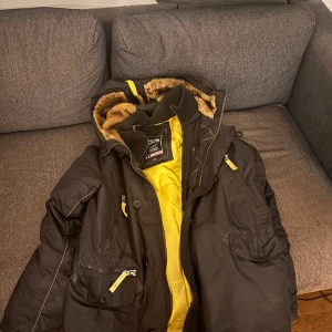 Alpha industries vinterjacka - Säljer en svart jacka från Alpha Industries med gul insida och pälsfodrad huva. Jackan har flera fickor med dragkedjor och knappar, perfekt för kalla dagar. Den är i storlek Large och har en cool, militärinspirerad stil.  Priset går att diskuteras