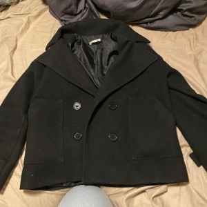 Svart kappa från Design - Säljer en stilren svart kappa från Design. Den har en klassisk dubbelknäppt stil med stora knappar och bred krage. Perfekt för höst och vinter, och den är i bra skick. Passar både till vardags och lite finare tillfällen.  Storlek xs/s💕