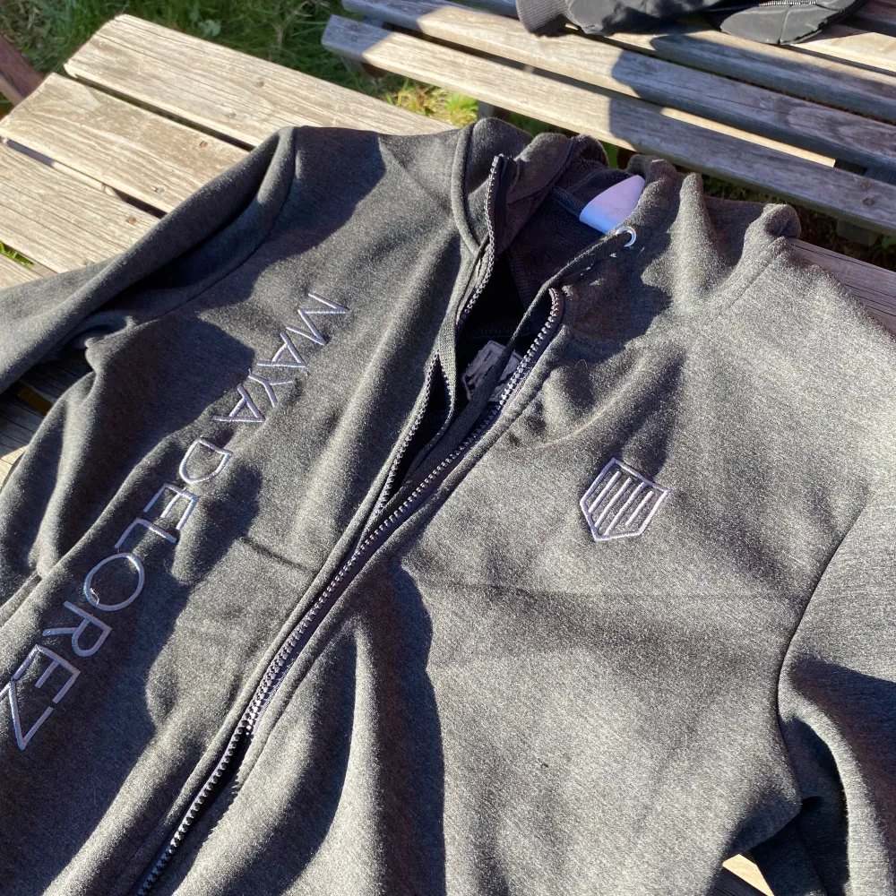 Säljer en snygg grå hoodie från Maya Delorez. Den har en dragkedja framtill och coola svarta detaljer i form av korsade band på ryggen. Perfekt för höst och vår, med långa ärmar och en bekväm passform. Märkesloggan syns både fram och bak. Perfekt för en casual look eller träning!. Hupparit & Collegepaidat.