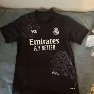 Säljer en snygg svart fotbollströja från Y-3 med Real Madrid-logga och 'Emirates Fly Better' tryck på framsidan. Tröjan har korta ärmar och ett coolt ros-mönster. Perfekt för alla fotbollsfans!